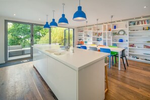 Et moderne køkken med en central ø, blå hængelamper og udsigt til grøn natur.