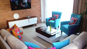 Ein Wohnzimmer mit Backsteinwand, Fernseher, Sofa, zwei Sesseln, Couchtisch und Schrank.
