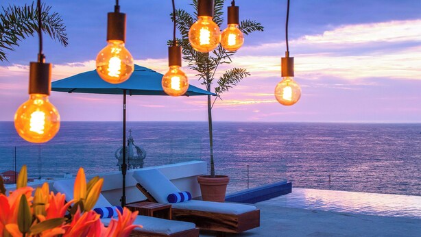 Eine Strandlounge mit hängenden Lampen, einem blauen Sonnenschirm und Blick auf den Ozean.