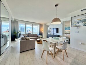 En moderne stue med sofa, spisebord og stole, et stort vindue med udsigt over byen, samt en loftslampe.
