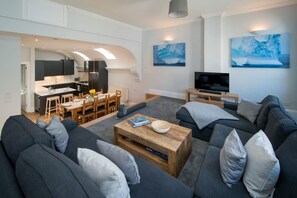 Uma sala de estar moderna com sofá, mesa de centro e televisão. Há uma área de jantar com cadeiras e uma mesa.