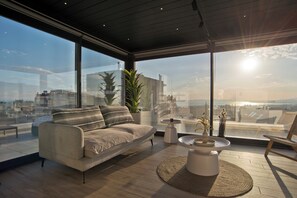 A modern living room with a sofa, a coffee table, and a view of the cityscape through large windows. - Riviera Sky Loft (Glifada)