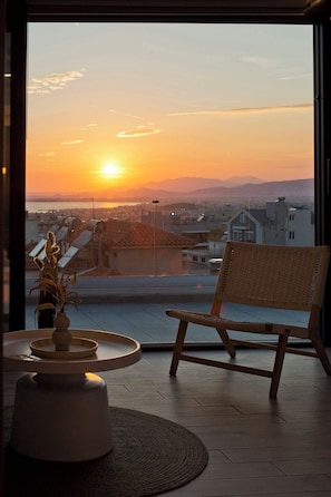 Ein Zimmer mit Blick auf einen Sonnenuntergang über einer Stadtlandschaft, ein Korbstuhl und ein kleiner Tisch mit einer Vase und einer Fernbedienung.