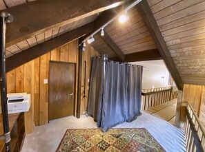 A room with a sloped ceiling, a wooden door, a window with curtains, a patterned rug, and a small table with a printer.