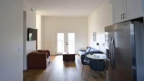 Eine moderne Küche mit einem Edelstahl-Kühlschrank, eine Essecke mit rundem Tisch und Stühlen, ein braunes Sofa und ein großes Fenster mit Blick.