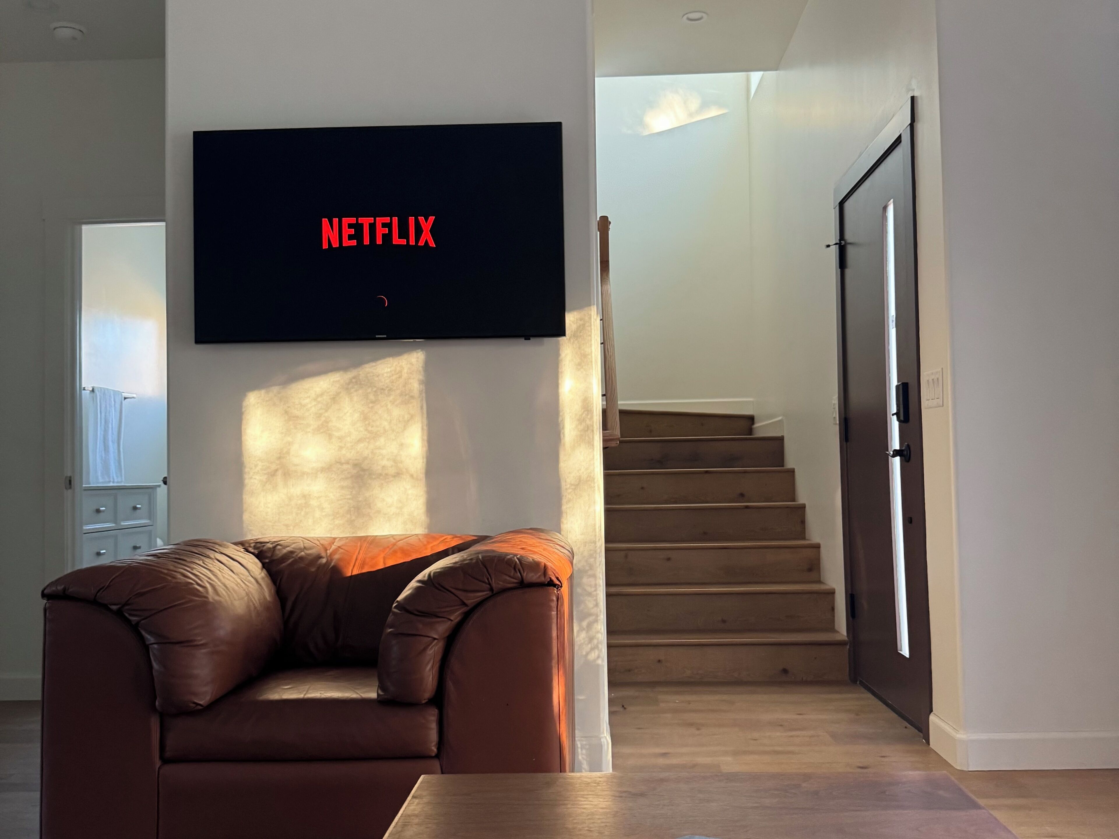 A living room with a brown leather sofa, a flat-screen TV mounted on the wall, and a staircase leading to another level.