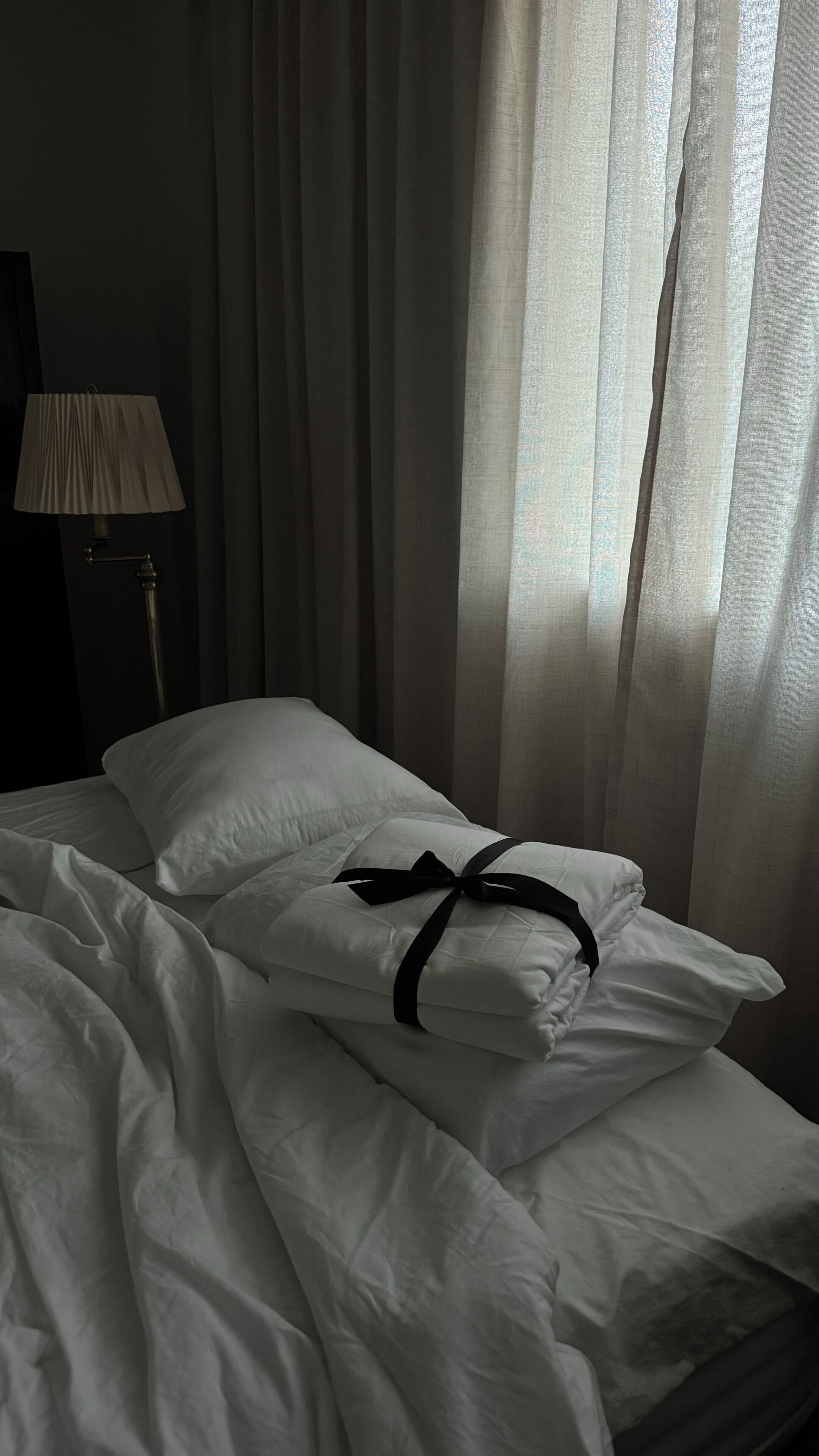 Un lit avec du linge de lit blanc et un ruban noir, une lampe et des rideaux en voile.