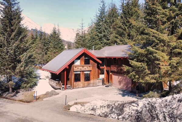 A wooden lodge with a sloped roof, a porch, and a garage, surrounded by trees and mountains.