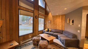 A cozy living room with a sofa, ottomans, a coffee table, and large windows. - Iconic A-Frame | Steps to Alyeska + Restaurants (Anchorage)