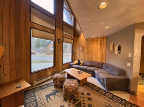A cozy living room with a sofa, ottomans, a coffee table, and large windows.
