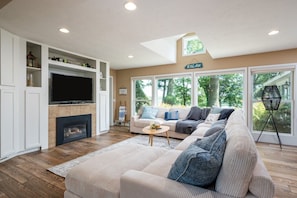 A living room with a sectional sofa, a flat-screen TV, a fireplace, and a large window with a view of trees. - Lakeside Retreat Mona Lake Waterfront Sleeps 12 (Norton Shores)