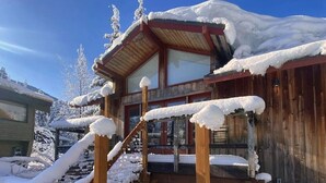A wooden cabin with a snow-covered roof, a wooden staircase, and a fenced area.