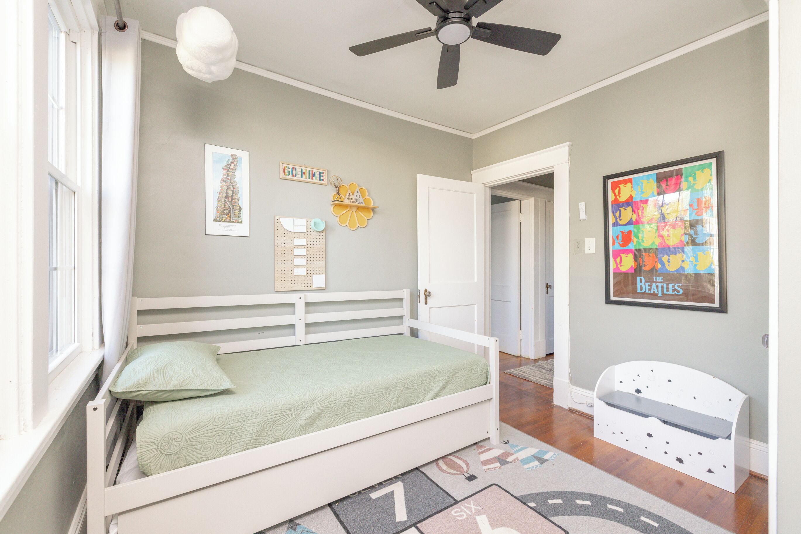 Un dormitorio con cama, ventilador de techo, ventana con cortinas, una puerta y una pared decorada con cuadros.