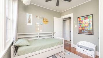 Un dormitorio con cama, ventilador de techo, ventana con cortinas, una puerta y una pared decorada con cuadros.
