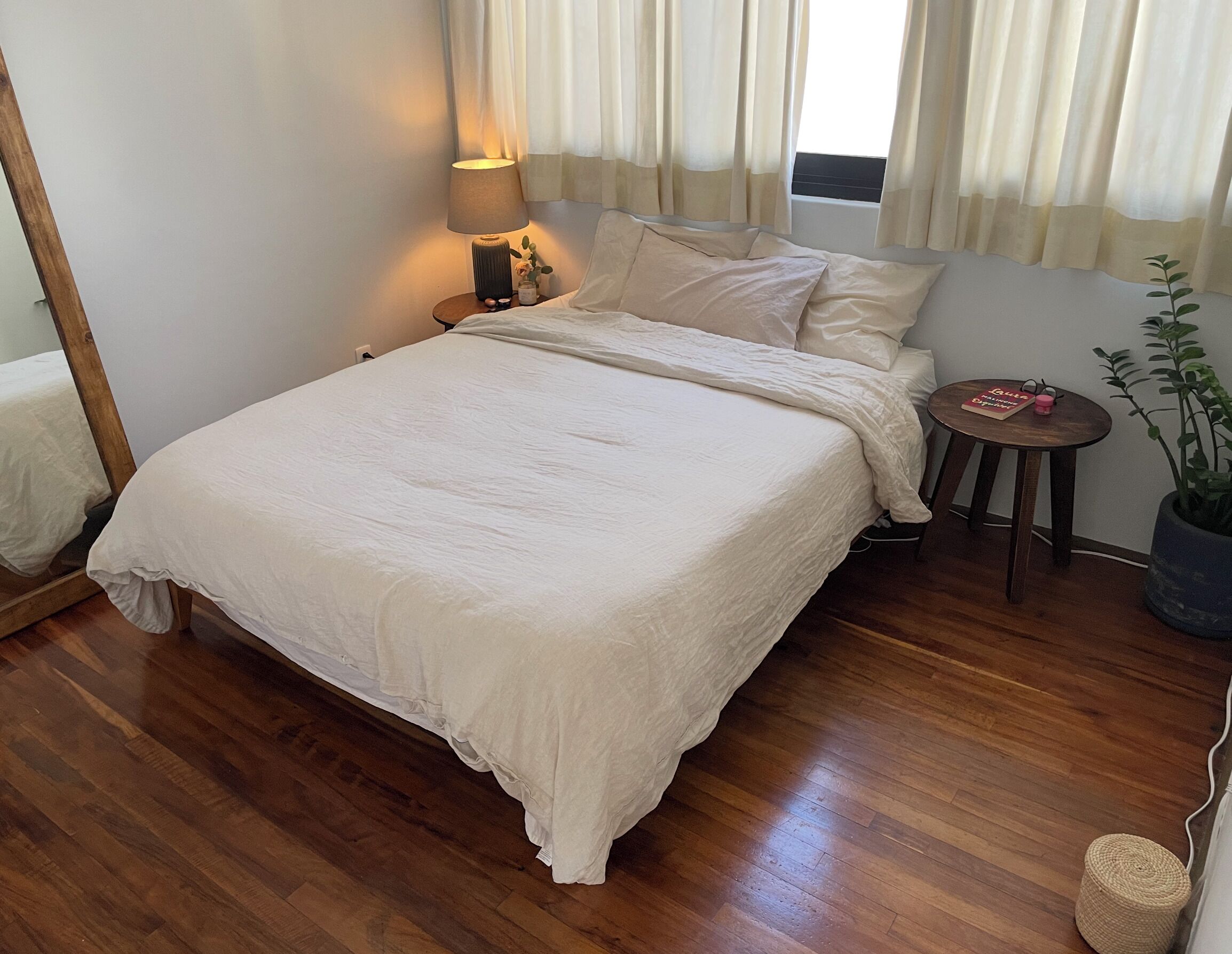 Una cama bien hecha con una colcha blanca y almohadas, una lámpara de noche, una pequeña mesa redonda con un libro y un objeto rosa, una planta en maceta y un suelo de madera.