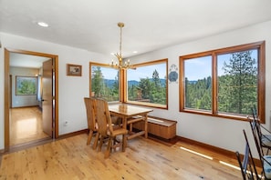 A dining area with a wooden table and chairs, large windows with a forest view, and a chandelier.