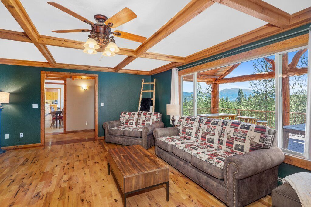 Une pièce dotée d’un plancher en bois, de deux canapés ornés de coussins à motifs, d’une table basse en bois, d’un ventilateur de plafond et de grandes fenêtres offrant une vue sur la verdure.