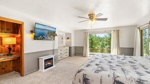Une chambre avec une cheminée, un ventilateur de plafond, un grand lit, une télévision et une vue sur les arbres par la fenêtre.
