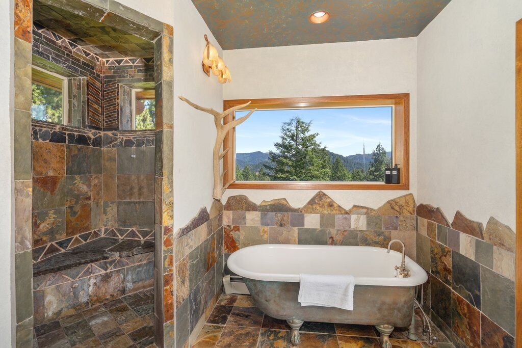 Une salle de bain avec une baignoire sur pieds, une fenêtre donnant sur un paysage de arbres et de montagnes, et une douche carrelée en pierre.