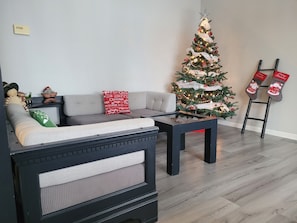 Ein Wohnzimmer mit grauem Sofa, einem Couchtisch, einem Weihnachtsbaum und einer Leiter mit Weihnachtsmützen.