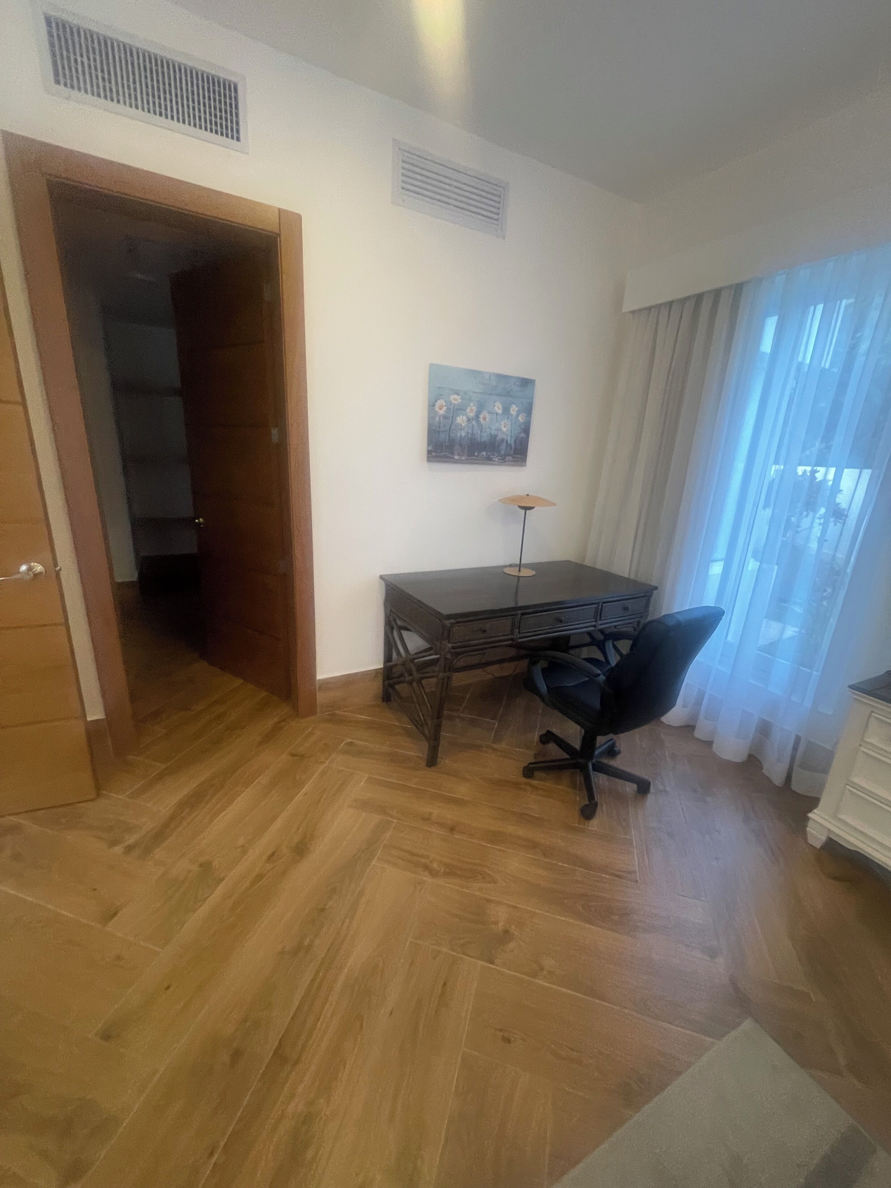 A room with a wooden floor, a desk with a chair, a painting on the wall, and a window with curtains.