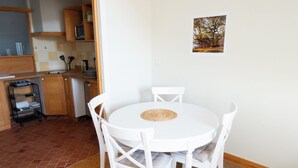 A white dining table with four chairs, a kitchen area with wooden cabinets, a microwave, and a small shelf with kitchen utensils.