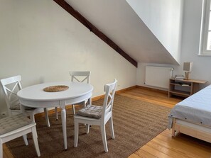 A room with a white table and chairs, a wooden floor, a bed with a striped blanket, a radiator, and a window.