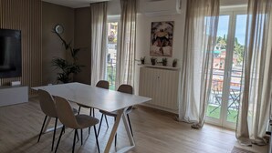 Ein modernes Wohnzimmer mit Esstisch, Stühlen, Fernseher, Pflanze und Blick ins Freie.