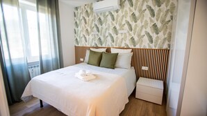Ein ordentlich bezogenes Bett mit weißer Decke und Kissen, ein Nachttisch mit Lampe, eine wandmontierte Klimaanlage und ein Fenster mit Vorhängen.