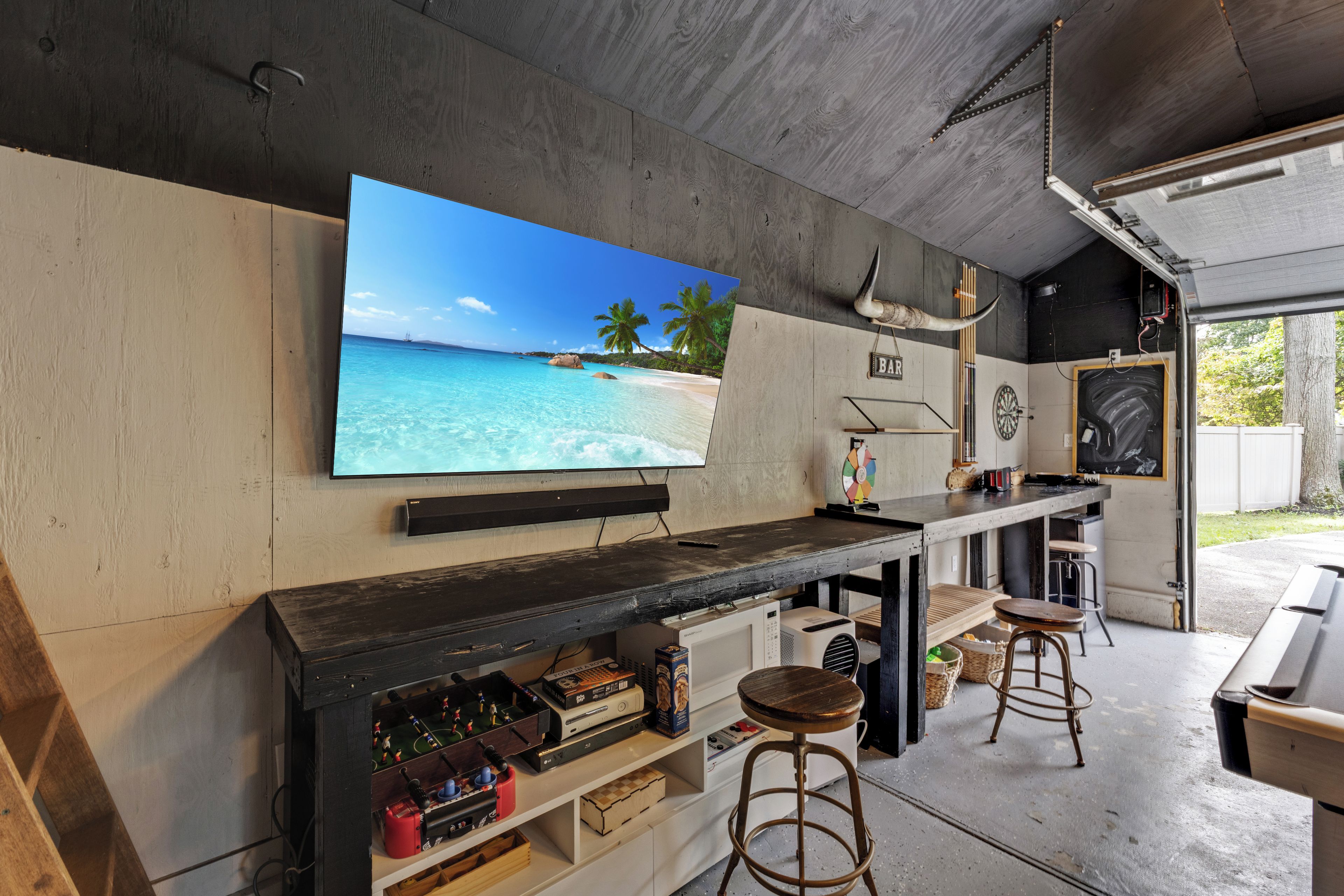 Zona de bar con una televisión mostrando una escena de playa, un dardo y estantes con diversos objetos.