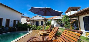 Uma área à beira da piscina com espreguiçadeiras de madeira e um guarda-sol, rodeada por edifícios com telhados em azulejo e paredes brancas.
