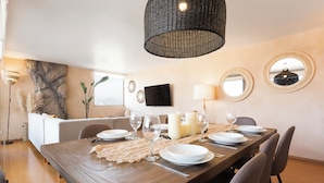 Un comedor con una mesa de madera dispuesta para cuatro, rodeada de sillas, un televisor montado en la pared y una lámpara de fibra trenzada grande colgada arriba.
