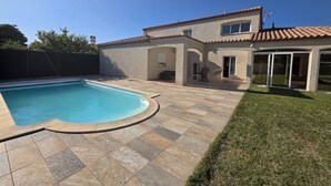 Une piscine avec une terrasse carrelée, un patio couvert et une maison avec un toit en tuiles.