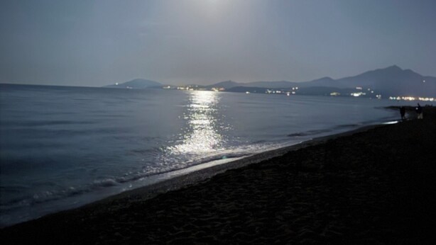 Une plage avec une mer calme, une lune bien visible et les lumières lointaines de la ville.