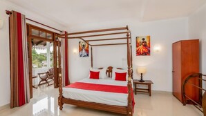 Una cama con dosel de madera, ropa de cama blanca y detalles rojos. Hay una silla de madera y una mesita de noche con una lámpara junto a la cama. Se observa un armario de madera en la esquina.