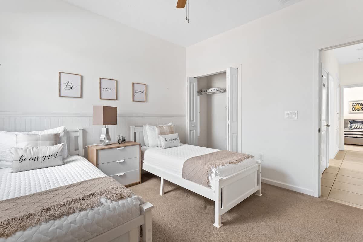 Une chambre avec deux lits, une table de chevet, une commode, une armoire et une porte donnant sur une autre pièce.