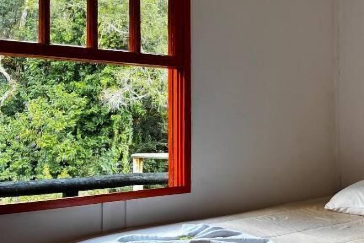 Una habitación con una cama, un ventanal de marcos rojos y vista a espacios verdes.