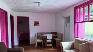 Une pièce avec un plancher en bois, un canapé marron, un canapé beige, une table à manger avec des chaises et une fenêtre avec des volets roses.