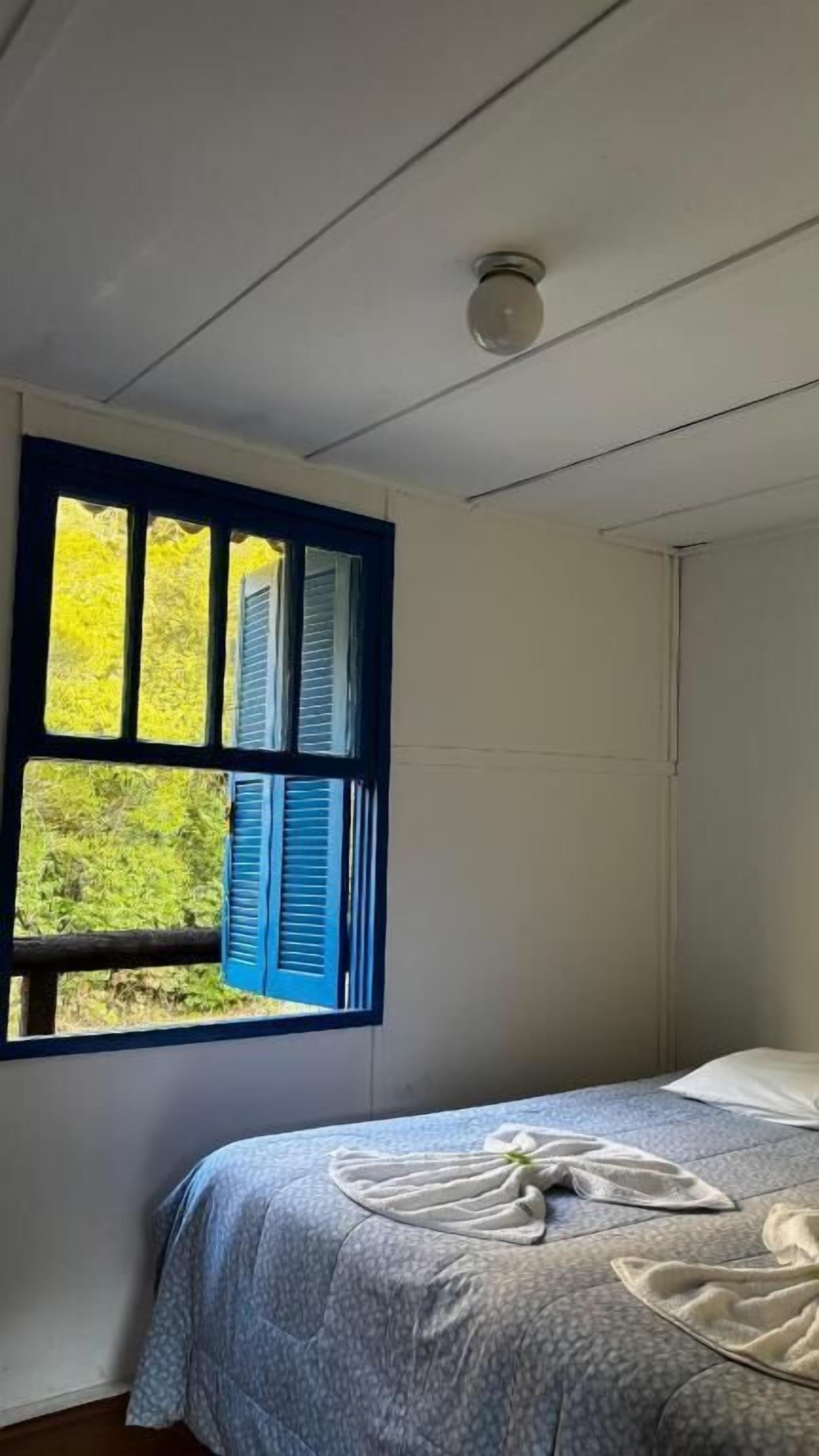 Un dormitorio con una cama, una ventana con persianas azules y vistas al verdor exterior.
