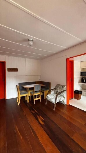 Una habitación con piso de madera, una mesa de comedor con sillas, un sillón estampado y una puerta roja.