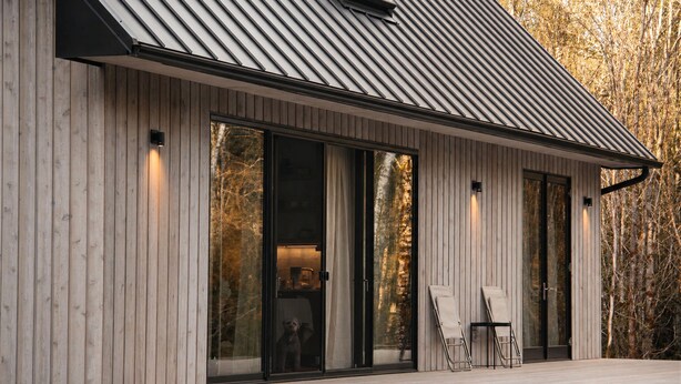 Un bâtiment moderne doté d’un toit en pente, de grandes portes vitrées et d’une terrasse en bois avec des chaises.