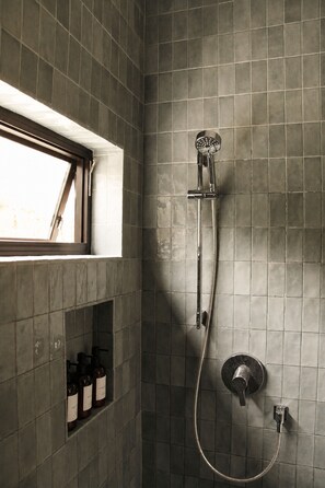 Eine Dusche mit Handbrause und Wandarmatur, ein Fenster mit Aussicht und ein Regal mit Flaschen.