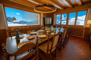 Een eetkamer met een grote houten tafel gedekt voor een maaltijd, grote ramen met uitzicht op een besneeuwde berg en een open haard.