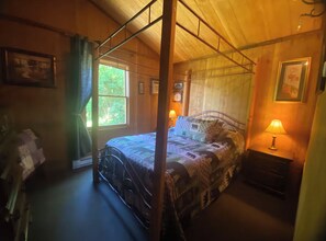 A bedroom with a four-poster bed, a nightstand with a lamp, a dresser, and a window with curtains.