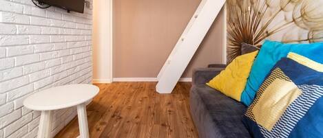 A room with a white brick wall, a mounted TV, a wooden floor, a sofa with patterned cushions, and a staircase.