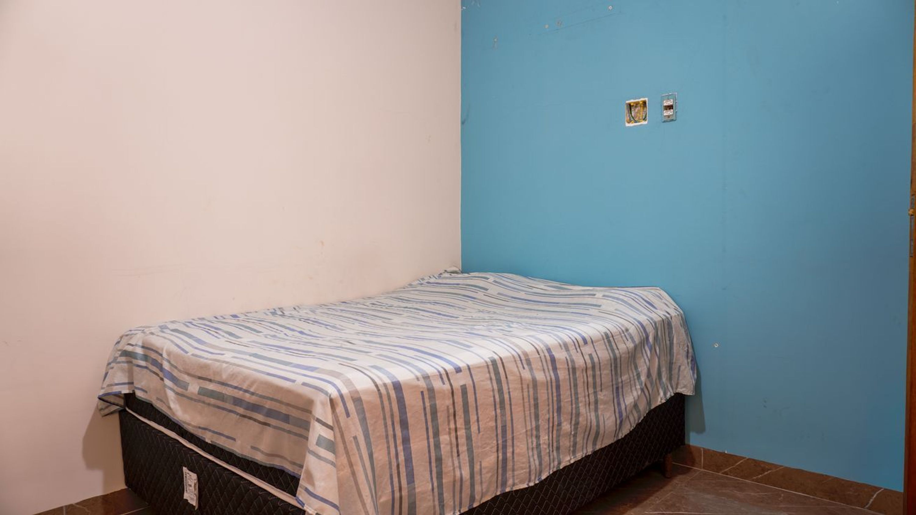 Una habitación pequeña con cama individual, colcha a rayas, colchón oscuro y pared azul.