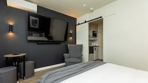 Ein modernes Hotelzimmer mit einem großen Bett, einem flachbildfernseher an der Wand, einem kleinen runden Tisch, einem grauen Sessel und einer Küchenzeile.