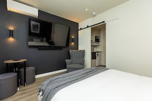 Ein modernes Hotelzimmer mit einem großen Bett, einem flachbildfernseher an der Wand, einem kleinen runden Tisch, einem grauen Sessel und einer Küchenzeile.