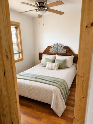 Une chambre à coucher avec un lit, une tête de lit en bois, un ventilateur de plafond et une fenêtre avec des stores.