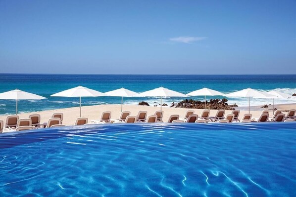Vista da piscina com espreguiçadeiras e guarda-sóis, com vista para a praia e o oceano.